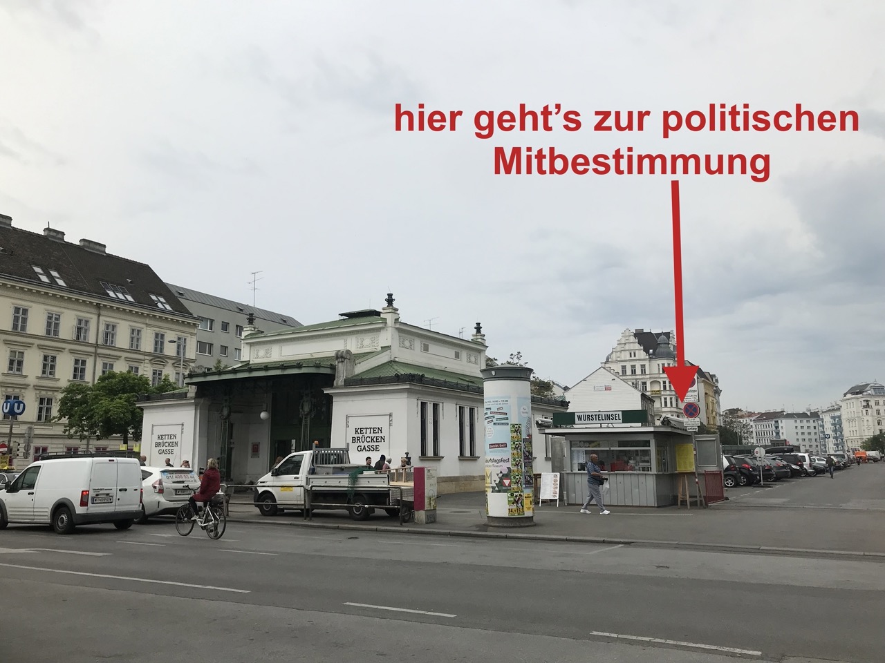 Fotografie Platz vor dem Stationsgebäude Kettenbrückengasse. Rechts im Bild ein kleiner, grauer Würstelstand. Hinweispfeil in Rot mit Beschriftung: hier geht's zur politischen Mitbestimmung zeigt auf bzw. hinter diesen Würstelstand.