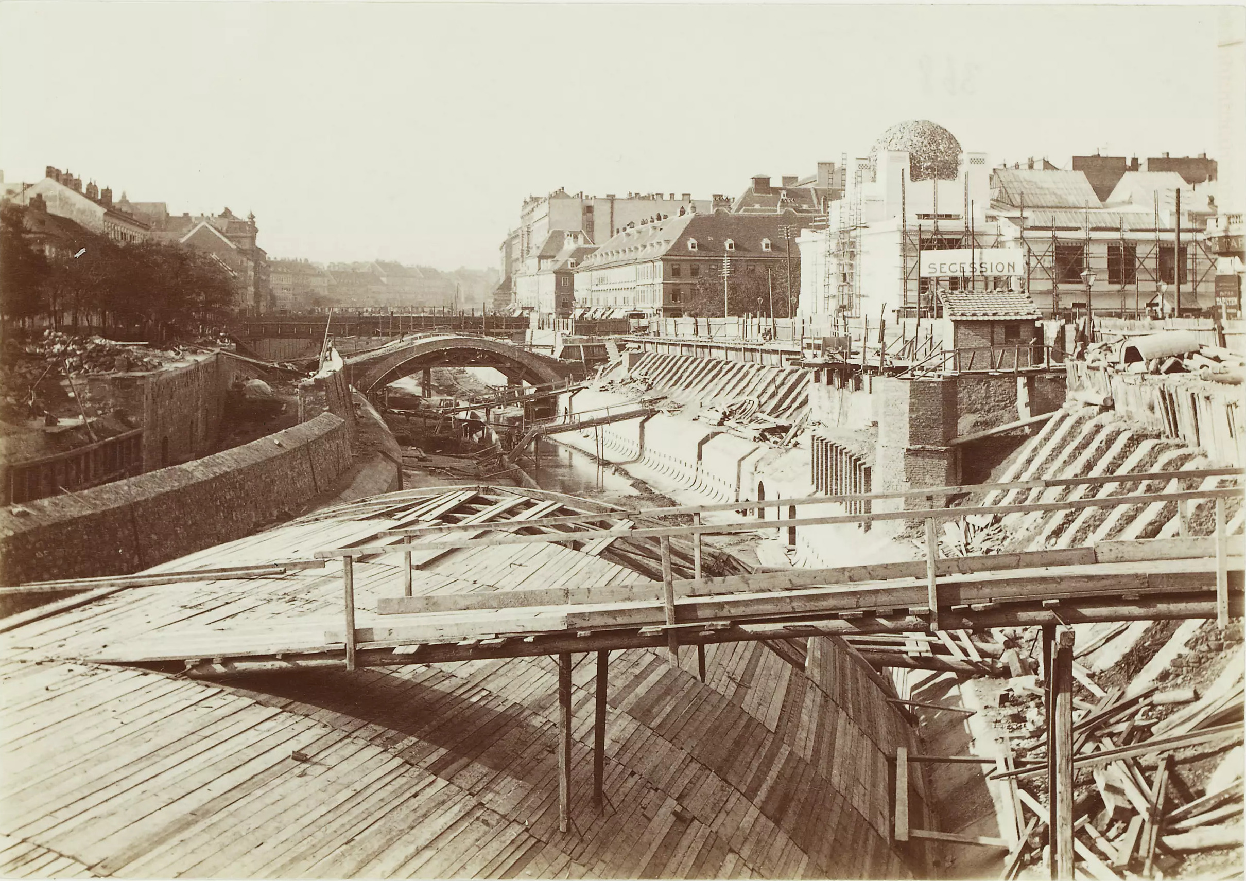 Schwarzweißfoto, sepia: Baustelle mit gewölbter Schalung aus Holz. Im Hintergrund stehen fertige Gebäude.