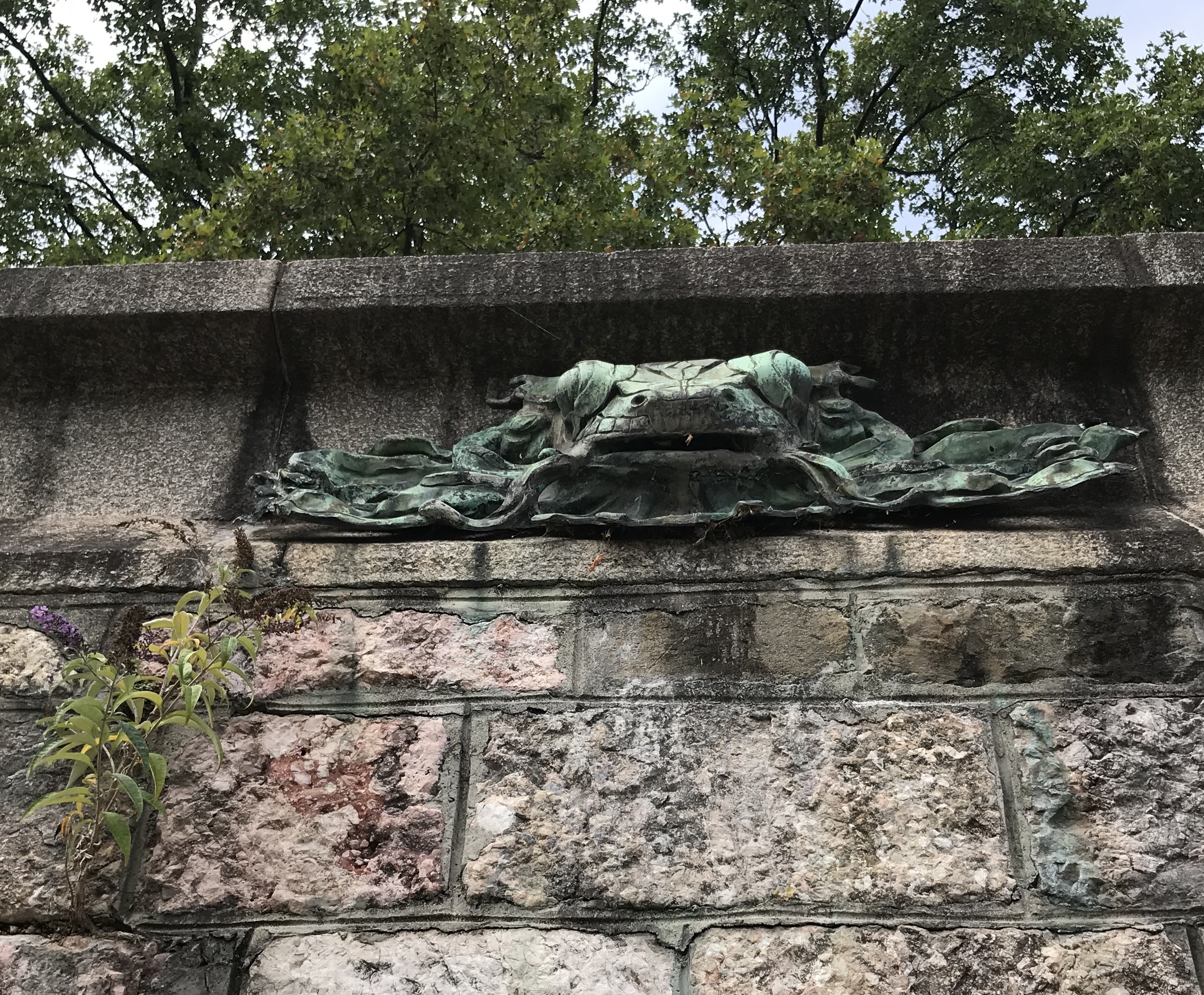 Farbfotografie: Mauerwerk aus rötlichen Steinquadern, aufgesetzt ein Froschkopf aus Bronze mit breitem Maul.