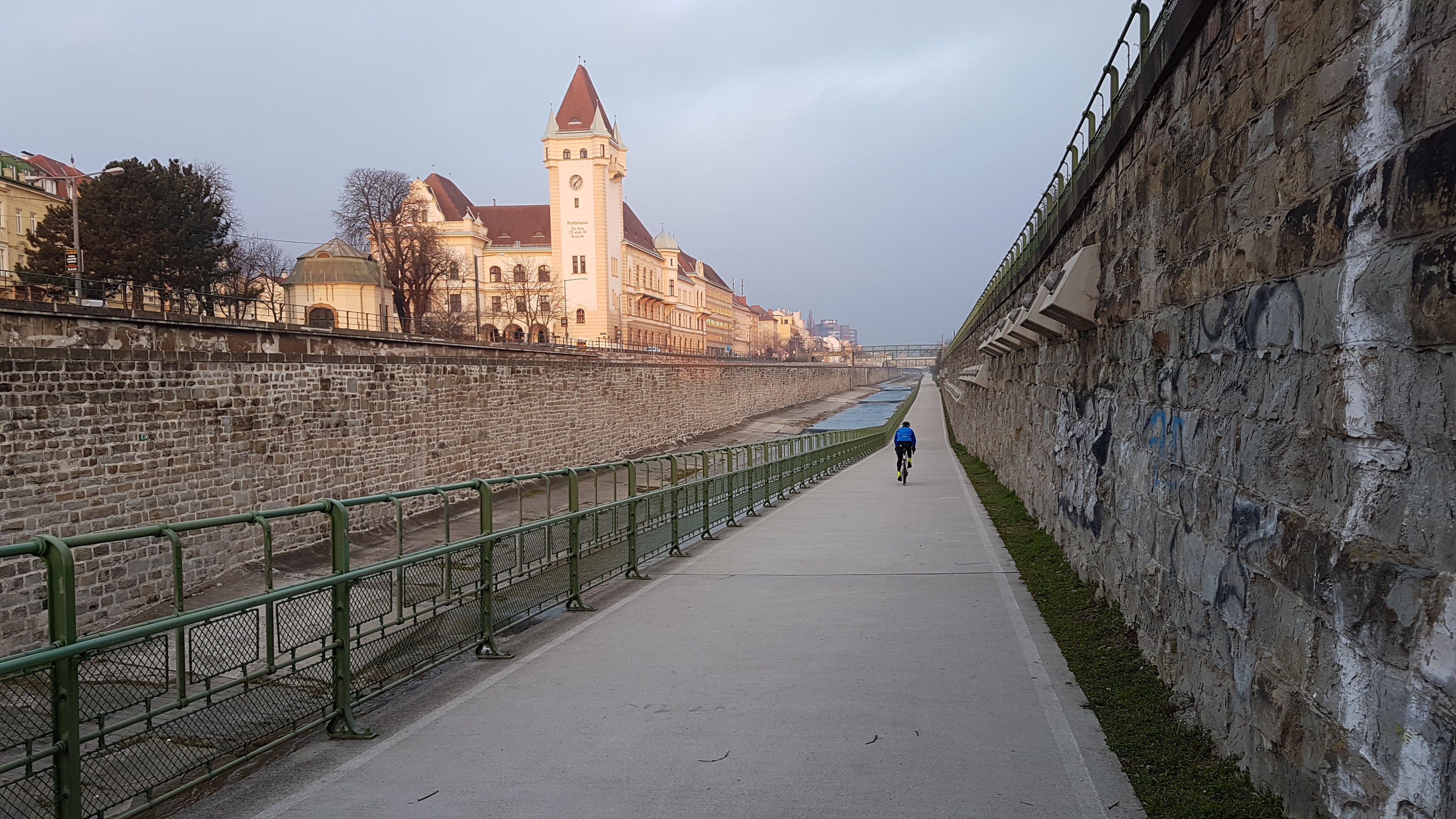 Eine Radwegrampe führt in ein mit hohen Steinmauren befestigtes Flussbett. Im Hintergrund ein schlossähnliches Gebäude in der Abendsonne.