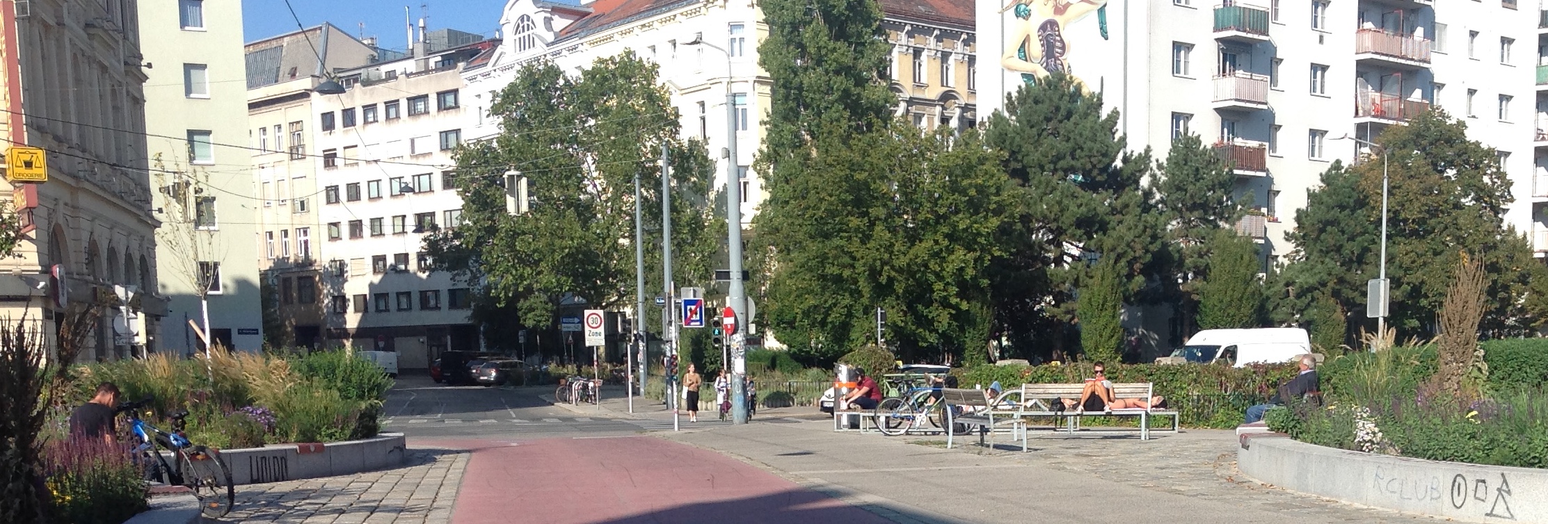 Menschen sitzen auf verschiedenen Sitzmöbeln in der Sonne. Im Hintergrund Bäume und Häuser.
