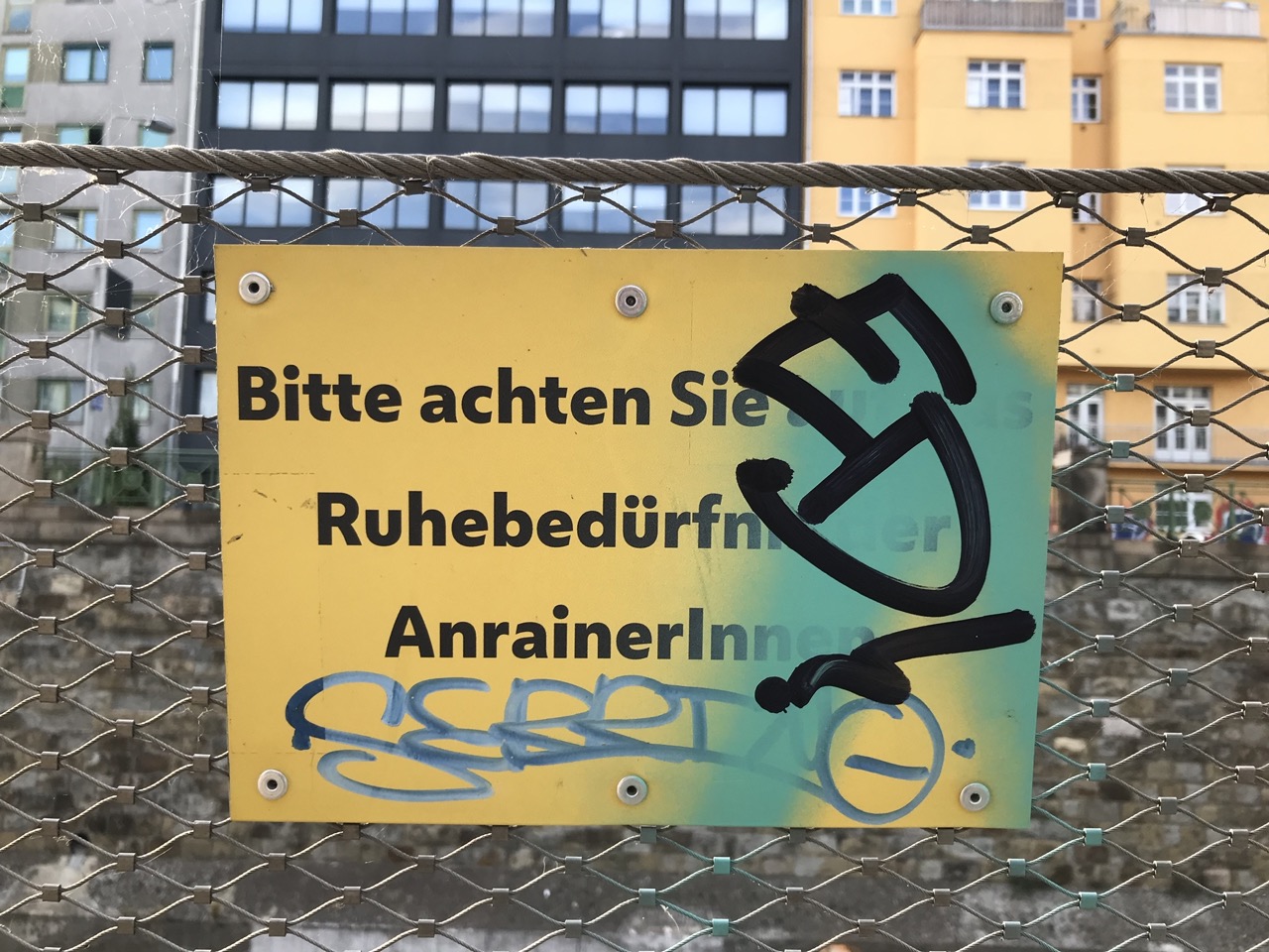 Schild mit Aufschrift: Bitte achten Sie auf die Ruhebedürfnisse der AnrainerInnen. Teilweise übermalt.