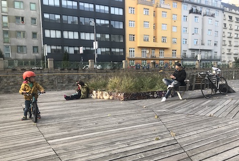 Kleinkind am Fahrrad, zwei Personen am Boden bzw. auf einer Bank rund um einen Pflanztrog mit Gräsern sitzend. Häuserzeile im Hintergrund.