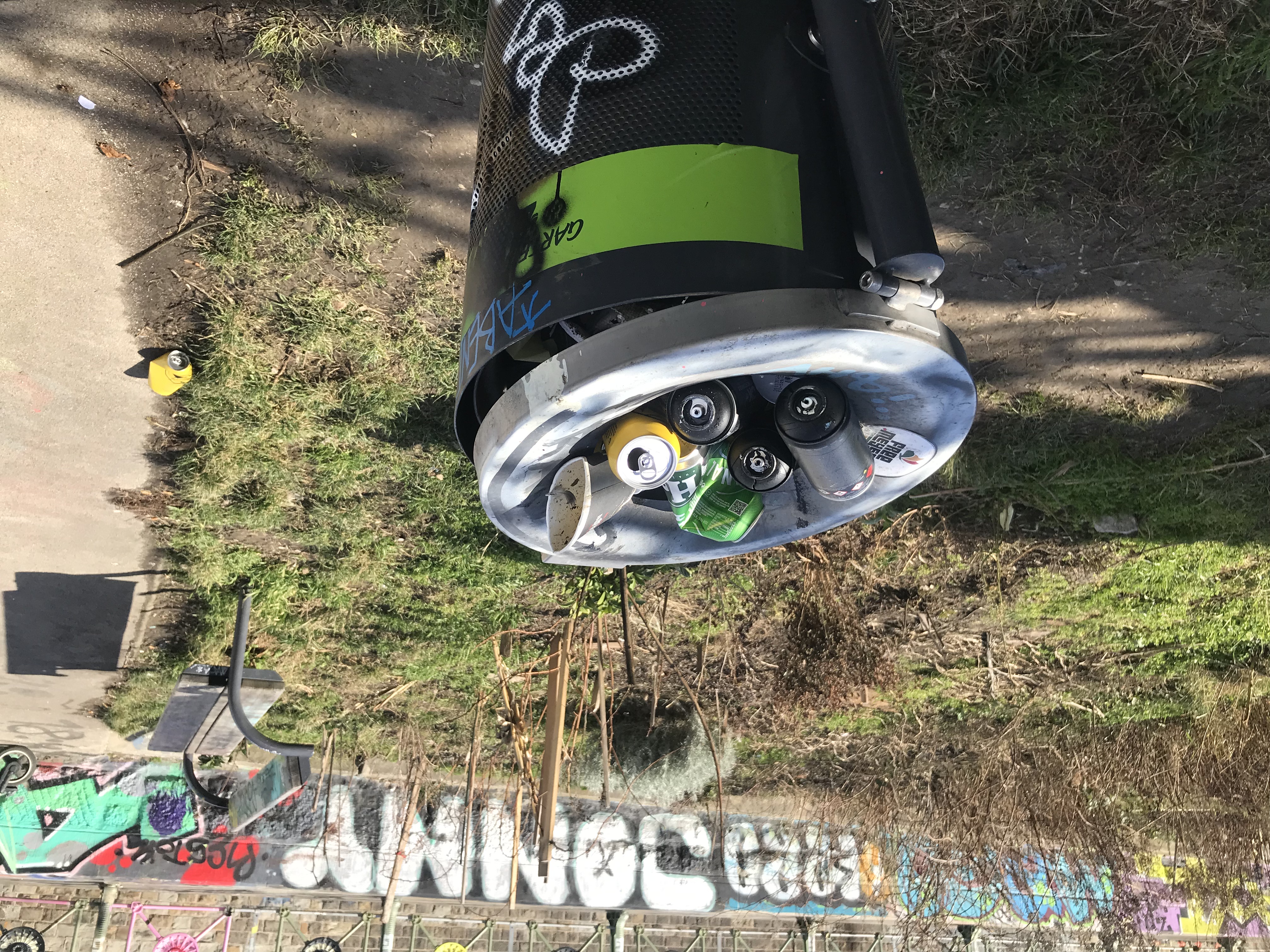 Mülleimer vor Sitzbank im Freien: Die Tonne ist randvoll, eine einzelne Bierdose liegt im Hintergrund am Boden. In der Tonne sind auch 3 Spraydosen zu sehen.