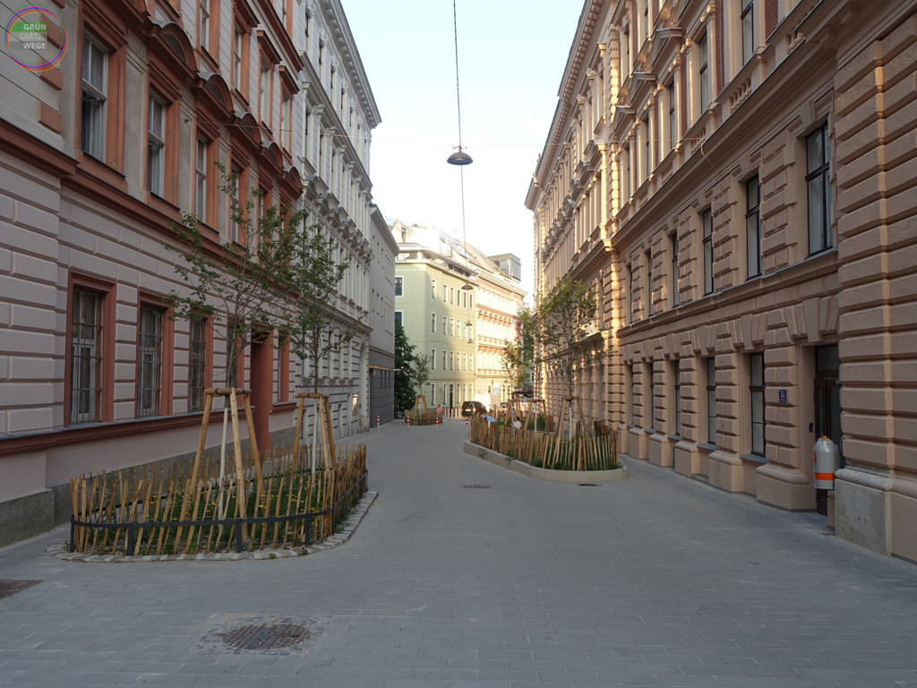 Schmale Straße in einem Gründerzeitblock als Fußgängerzone mit Grüninseln, Bäumen, im Hintergrund sitzen Menschen, in der Mitte verhindern zusätzlich zur gepflasterten Gestaltung auch Poller die Kfz-Durchfahrt
